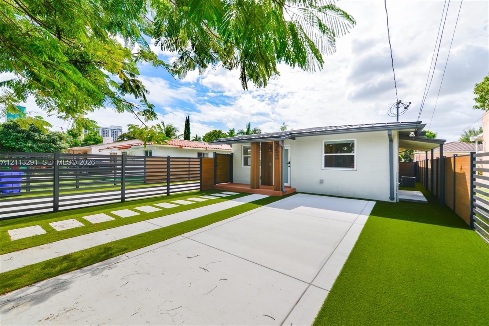 Exterior featuring light-colored masonry, horizontal slat fencing, a concrete driveway, paver stepping stones, and synthetic turf lawn Exterior featuring light-colored masonry, horizontal slat fencing, a concrete driveway, paver stepping stones, and synthetic turf lawn