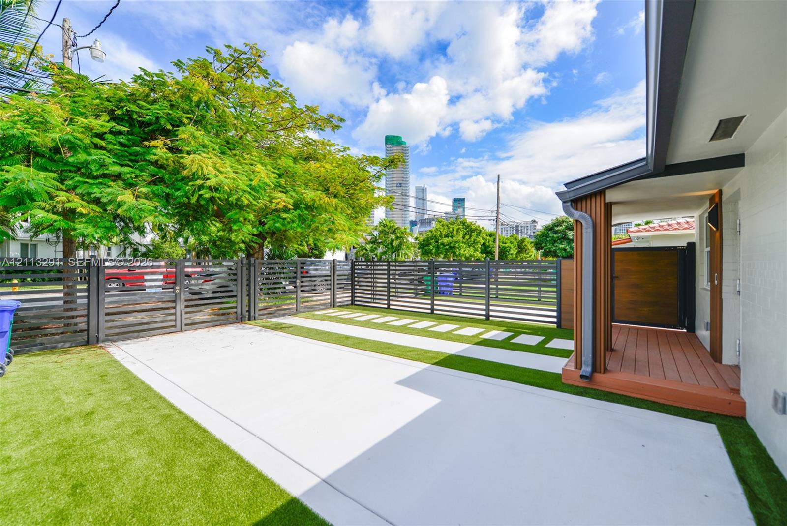 Fenced exterior space featuring concrete paving, synthetic turf, wood-finish decking, and slat-style metal fencing Fenced exterior space featuring concrete paving, synthetic turf, wood-finish decking, and slat-style metal fencing