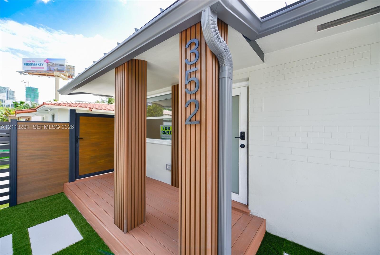 Entrance featuring a composite deck, vertical wood-slat accent columns, white painted brick exterior, and a privacy fence with a matching wood-finish gate Entrance featuring a composite deck, vertical wood-slat accent columns, white painted brick exterior, and a privacy fence with a matching wood-finish gate