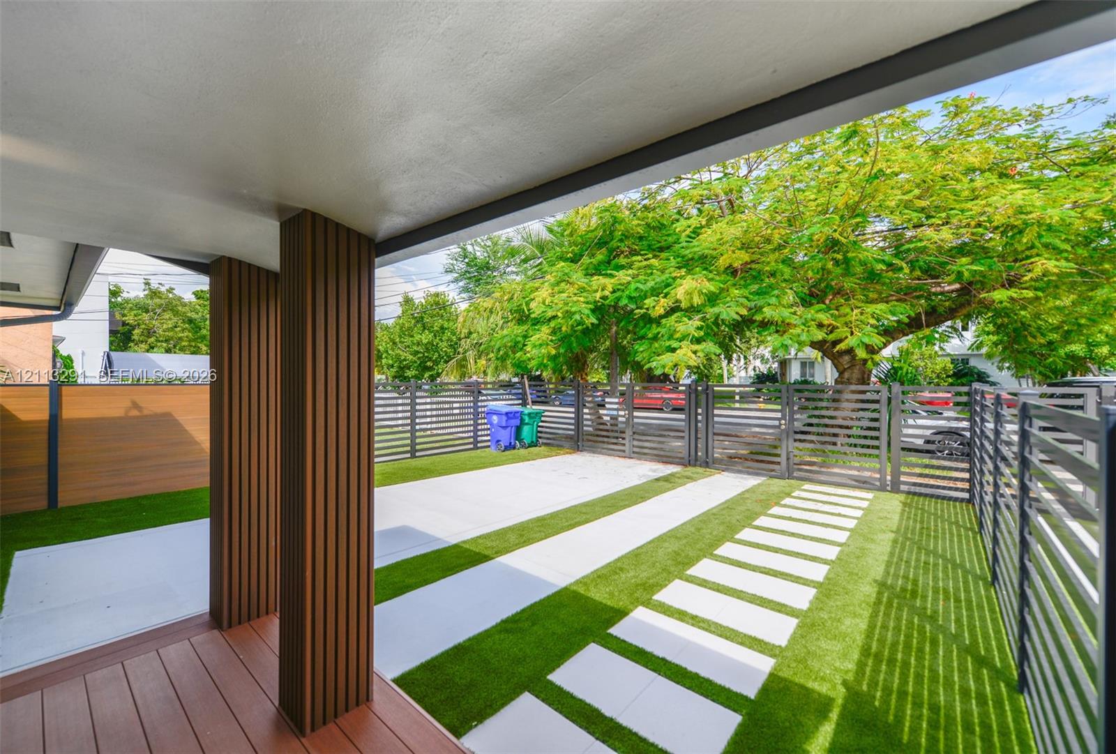 Covered patio featuring wood-finish decking and vertical slat columns, accompanied by a landscaped area with synthetic turf, concrete pavers, and horizontal metal privacy fencing