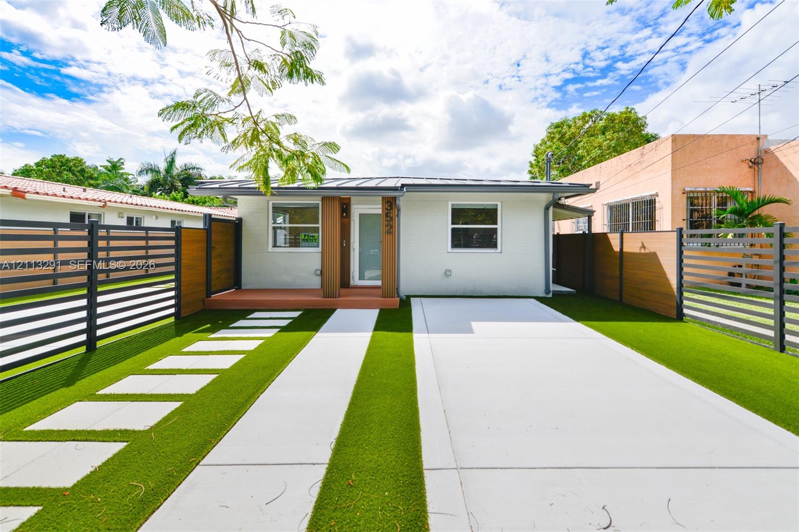 Single-story exterior featuring a metal roof, concrete driveway, synthetic turf lawn, stone paver walkway, and horizontal slat privacy fencing Single-story exterior featuring a metal roof, concrete driveway, synthetic turf lawn, stone paver walkway, and horizontal slat privacy fencing
