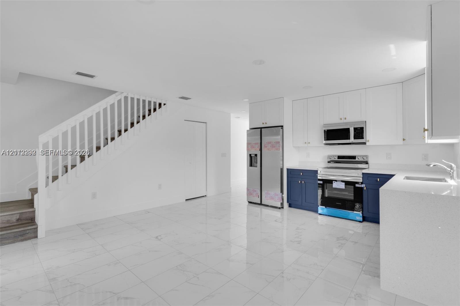Kitchen featuring white upper cabinetry, navy blue lower cabinetry, stainless steel appliances, and quartz countertops, complemented by white polished stone-finish flooring and a staircase with white wood railing