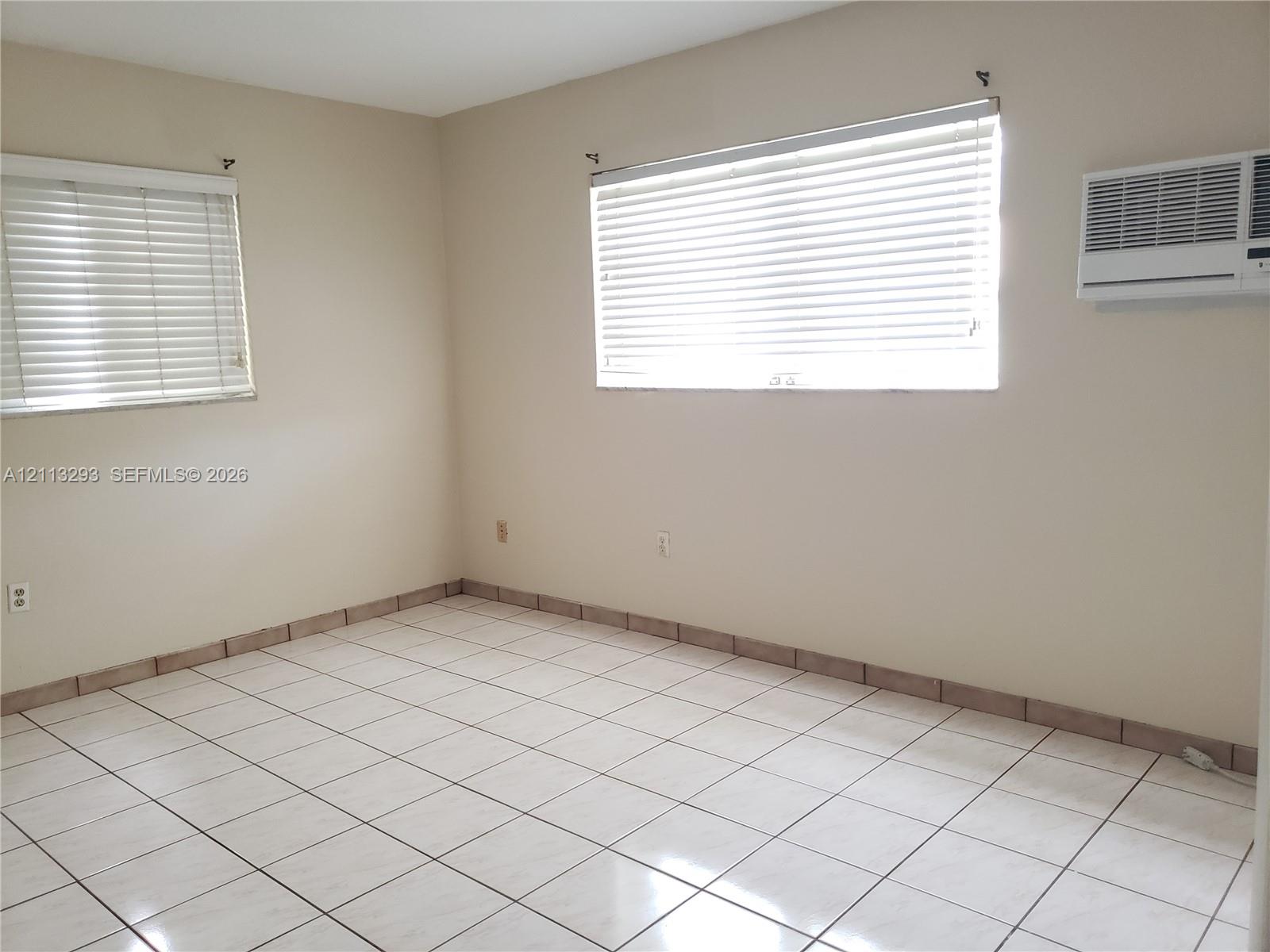 Interior space featuring neutral wall paint, white tile flooring, dual windows with fitted horizontal blinds, and a wall-mounted air conditioning unit