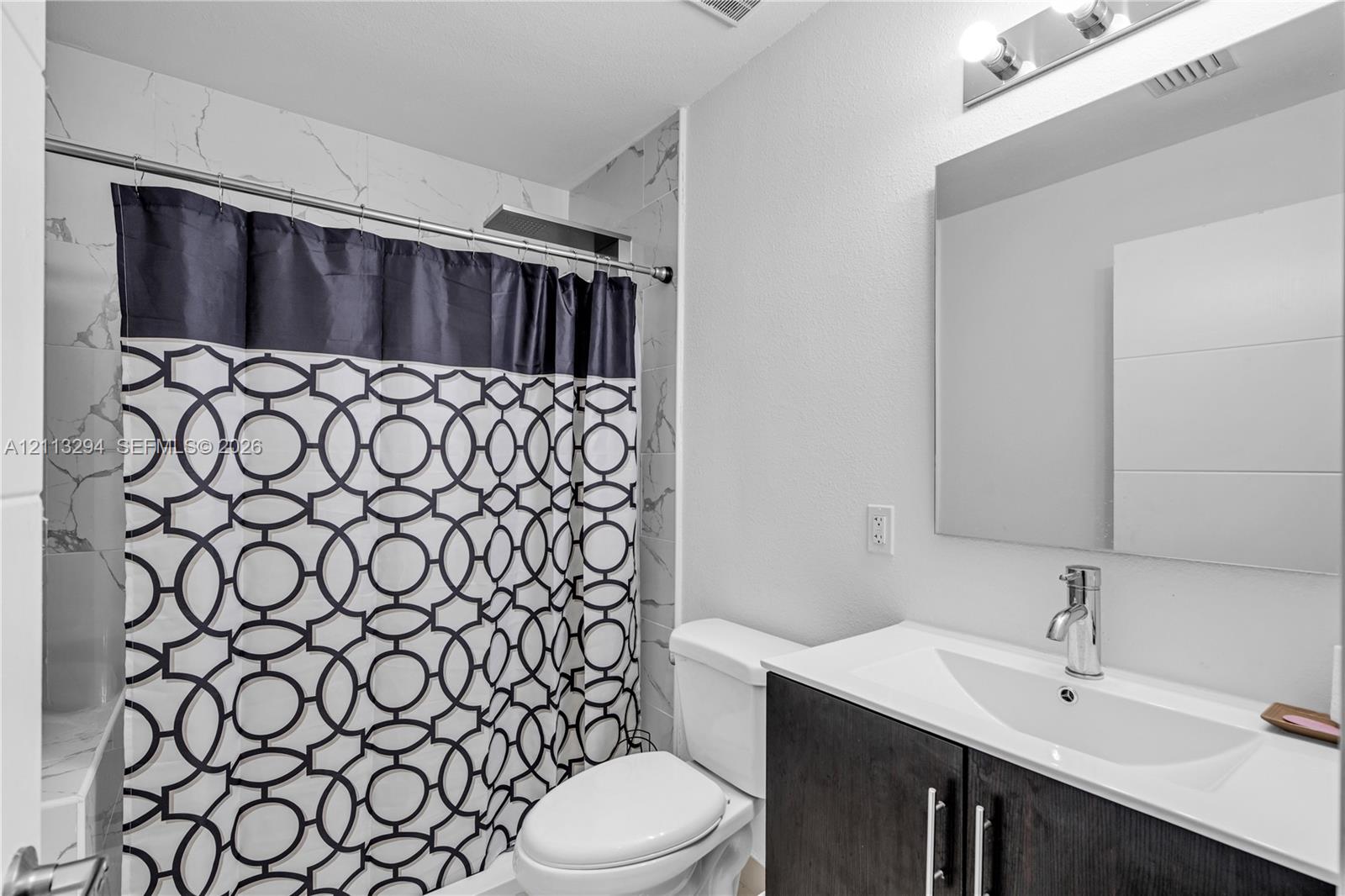 Bathroom featuring a dark-finish vanity with a rectangular sink, stone-pattern wall tile surround in the shower, polished chrome faucet, and a wall-mounted mirror with overhead lighting