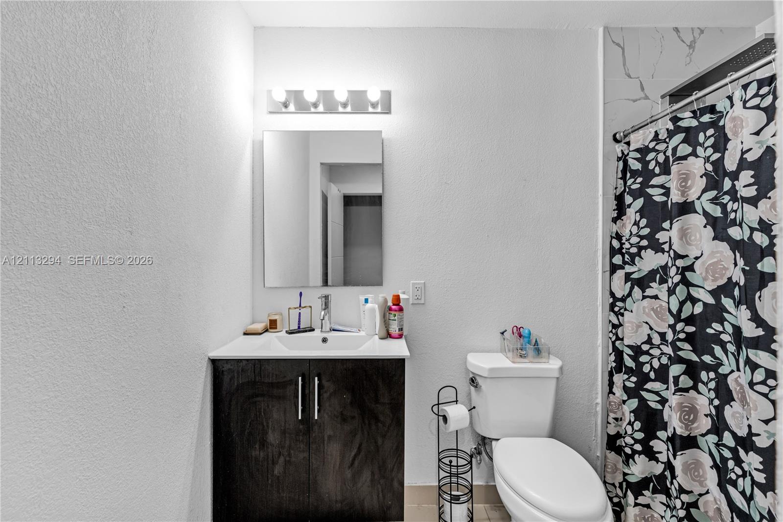 Bathroom featuring a vanity with dark wood-finish cabinetry, a white integrated sink, a rectangular wall-mounted mirror, a four-bulb light fixture, and a tiled shower surround