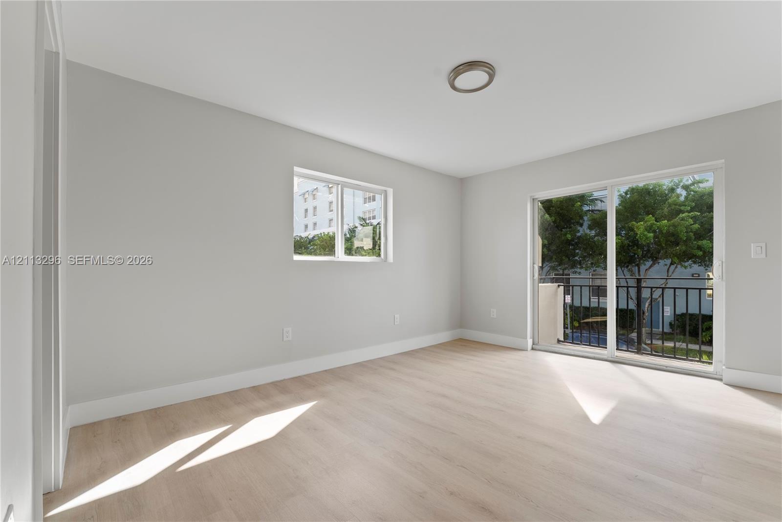 Neutral-toned interior featuring wood-finish flooring, a flush-mount light fixture, and sliding glass doors opening to a balcony with metal railing
