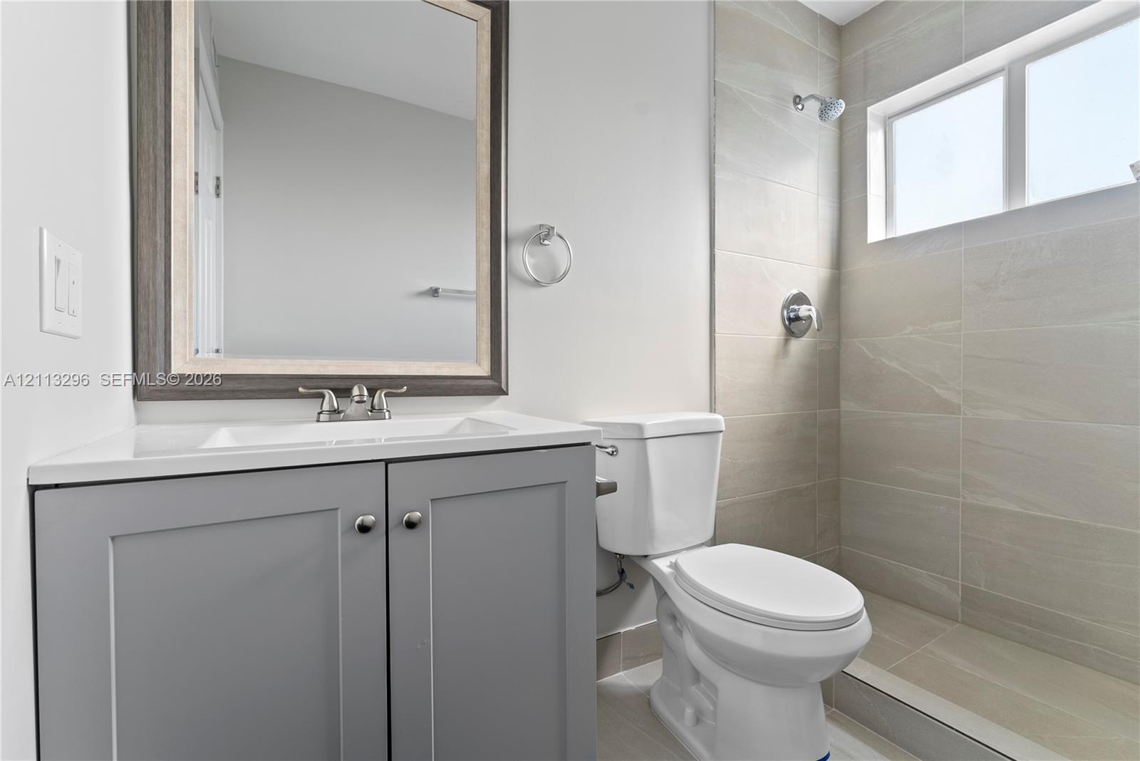 Bathroom featuring a gray shaker-style vanity with an integrated sink, framed wall-mounted mirror, ceramic tile shower surround, and a horizontal window