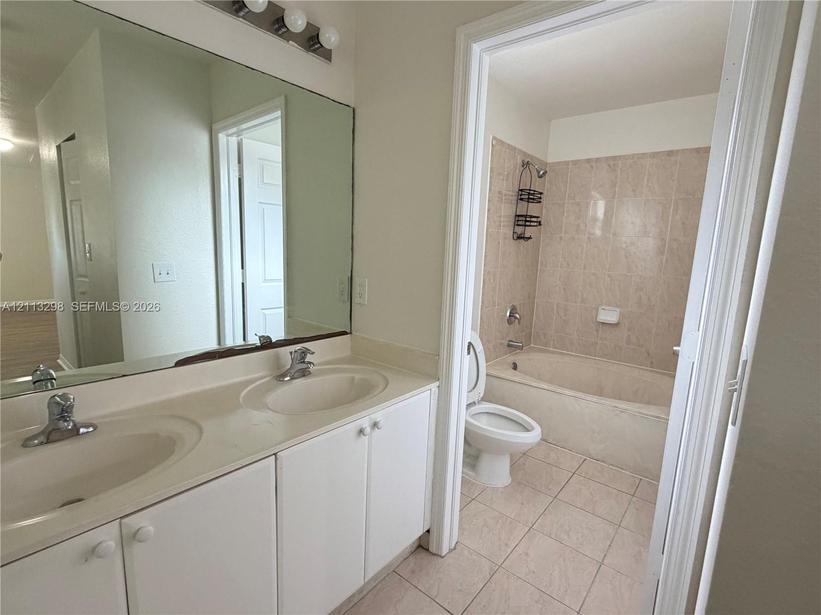 Dual-basin vanity with integrated sinks, large wall-mounted mirror, light-tone cabinetry, tiled flooring, and a combined tub and shower enclosure with wall-mounted ceramic tile surround