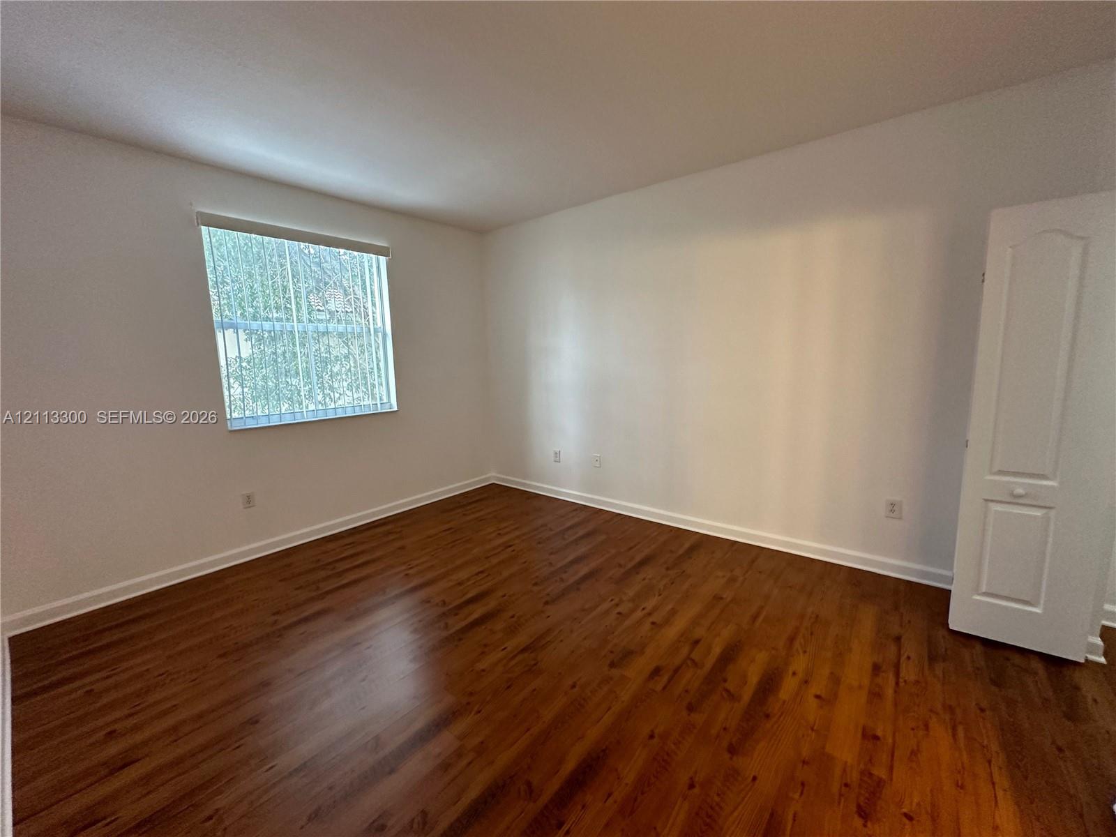 Room featuring wood-finish flooring, neutral walls, a single-panel interior door, and a window with vertical blinds