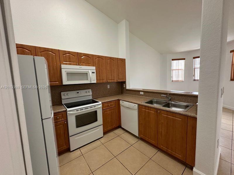 Kitchen featuring wood-finish cabinetry, neutral-toned countertops, double-basin stainless steel sink, ceramic tile flooring, and vaulted ceilings