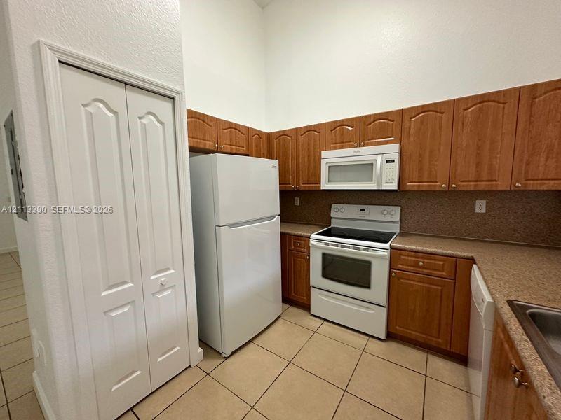 Kitchen featuring wood-finish cabinetry, white appliances, solid-surface countertops, tile flooring, and a bi-fold pantry door