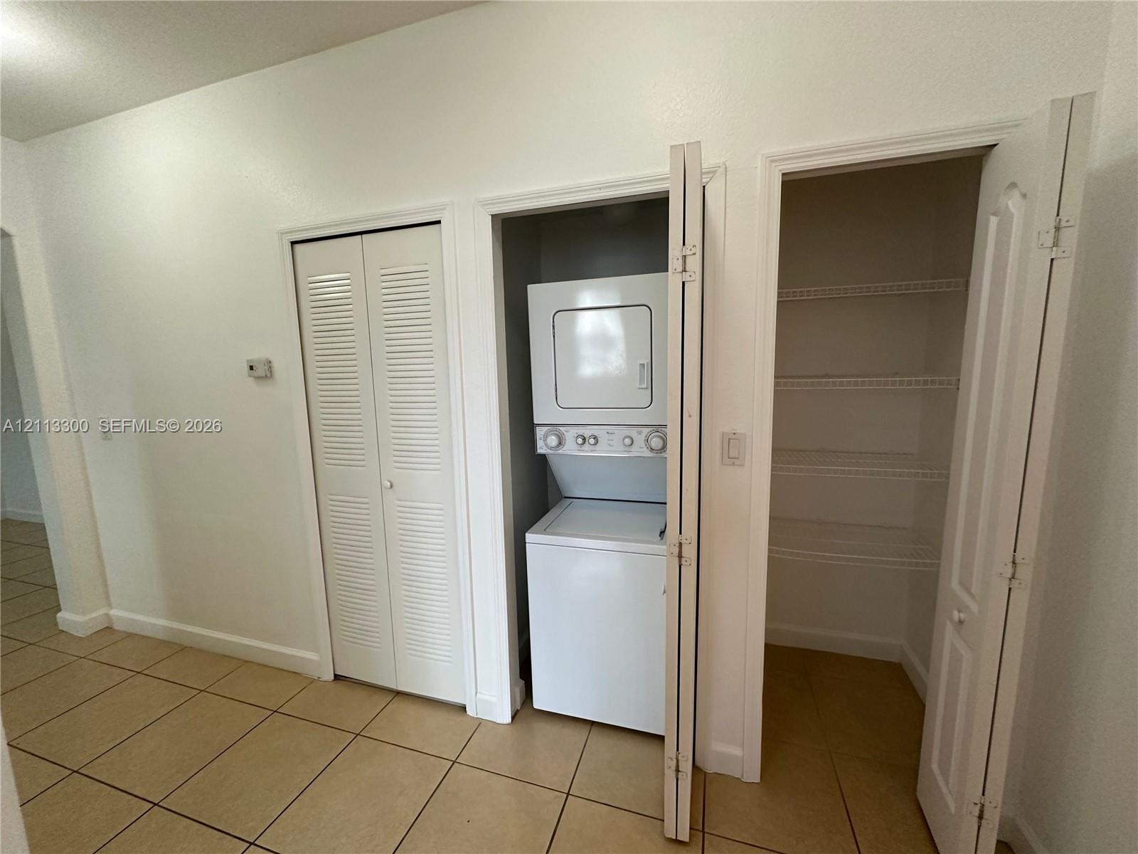 Utility area featuring neutral tile flooring, stacked washer and dryer unit, louvered closet doors, and built-in wire shelving