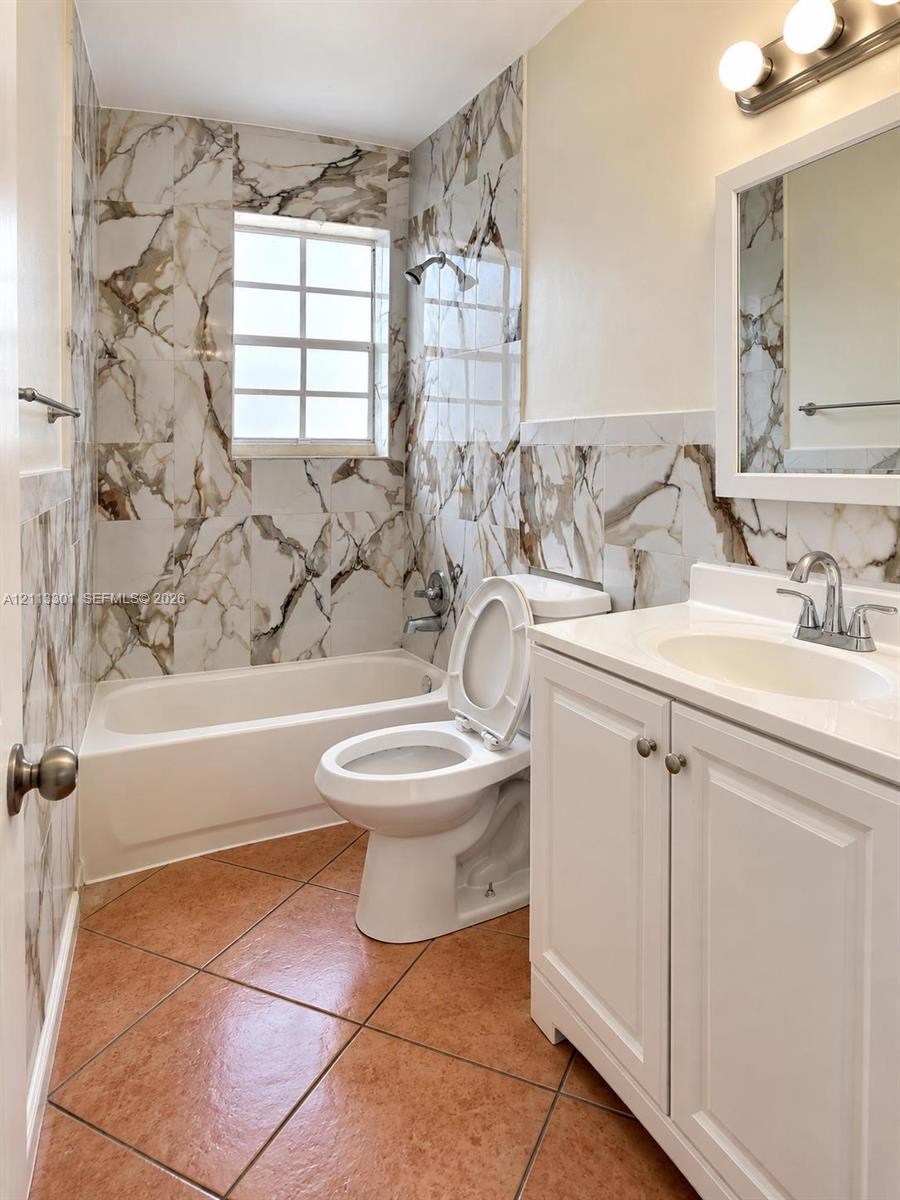 Bathroom featuring large-format patterned wall tile, white vanity cabinet with integrated sink and countertop, ceramic tile flooring, built-in tub with shower fixture, and framed wall mirror