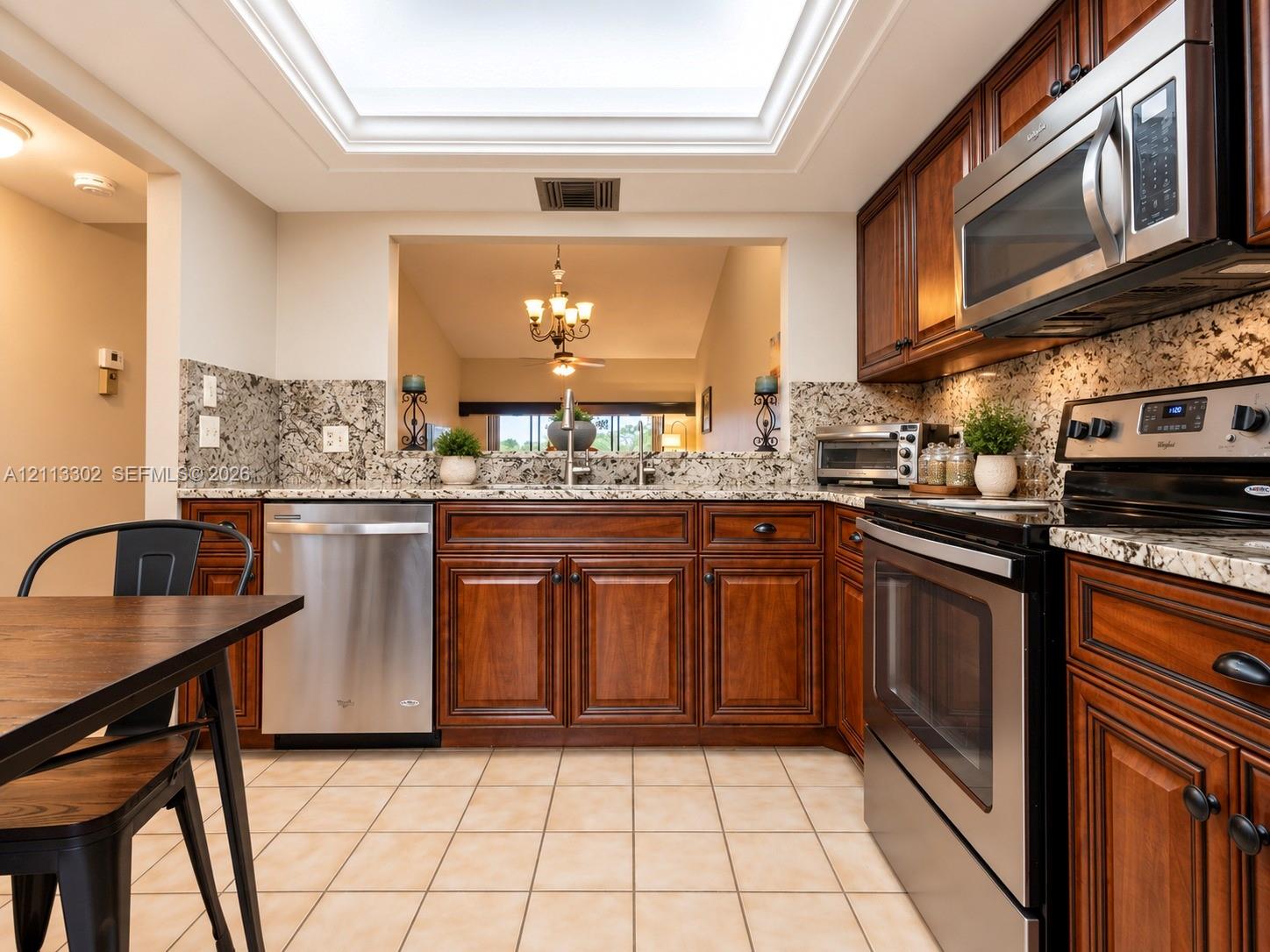 Kitchen featuring custom stained wood cabinetry, stone countertops, stainless steel appliances, tiled flooring, and a recessed ceiling skylight