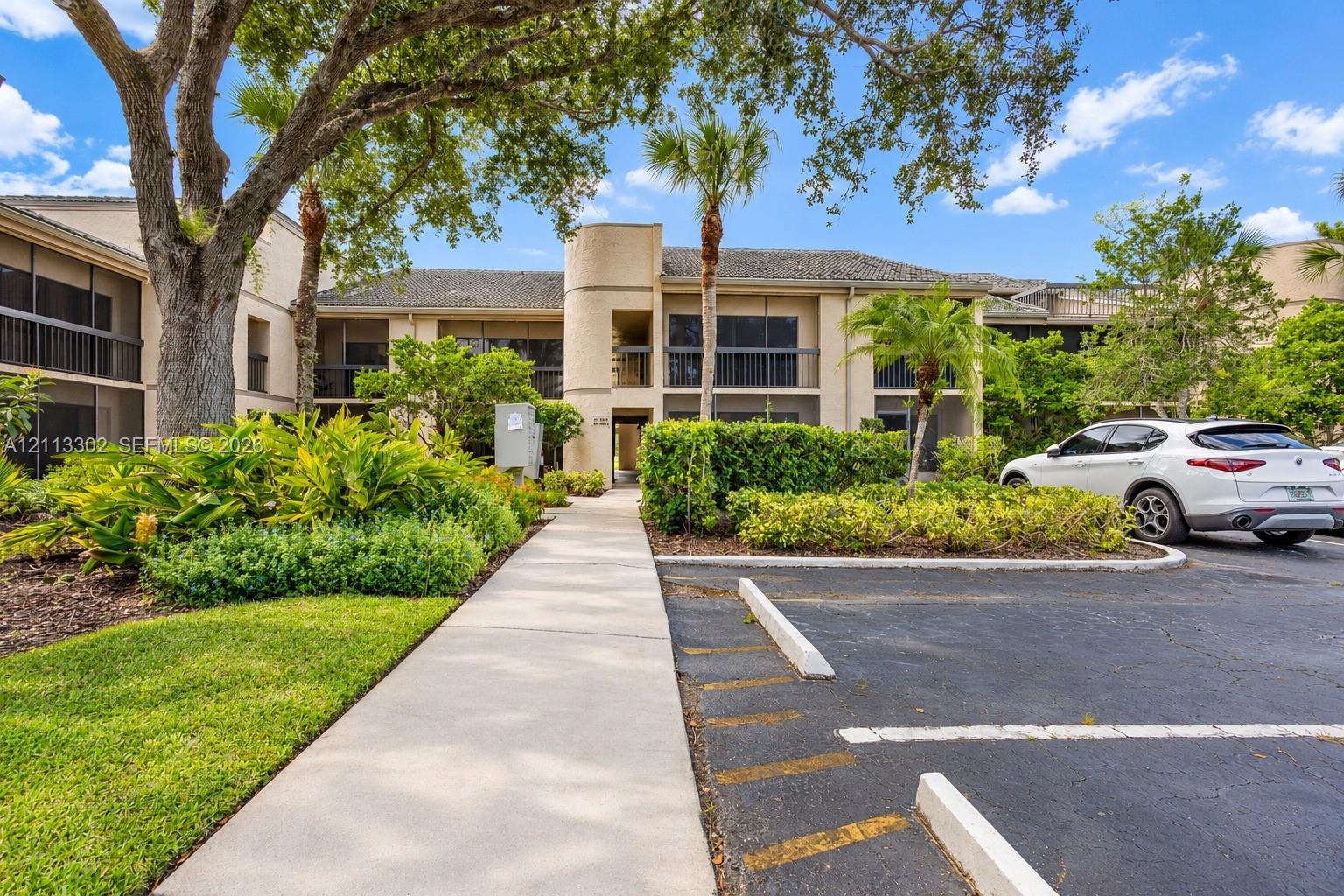 Multi-story exterior featuring a neutral stucco facade, screened balconies, mature shade trees, and manicured landscaping