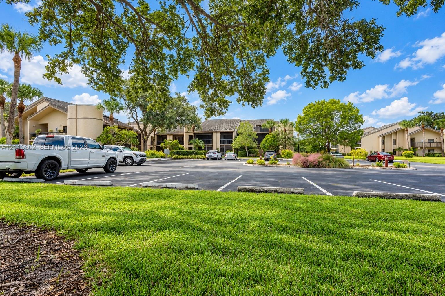 Exterior grounds featuring paved parking, expansive green space, mature canopy trees, and landscaped shrubbery surrounding multi-story buildings