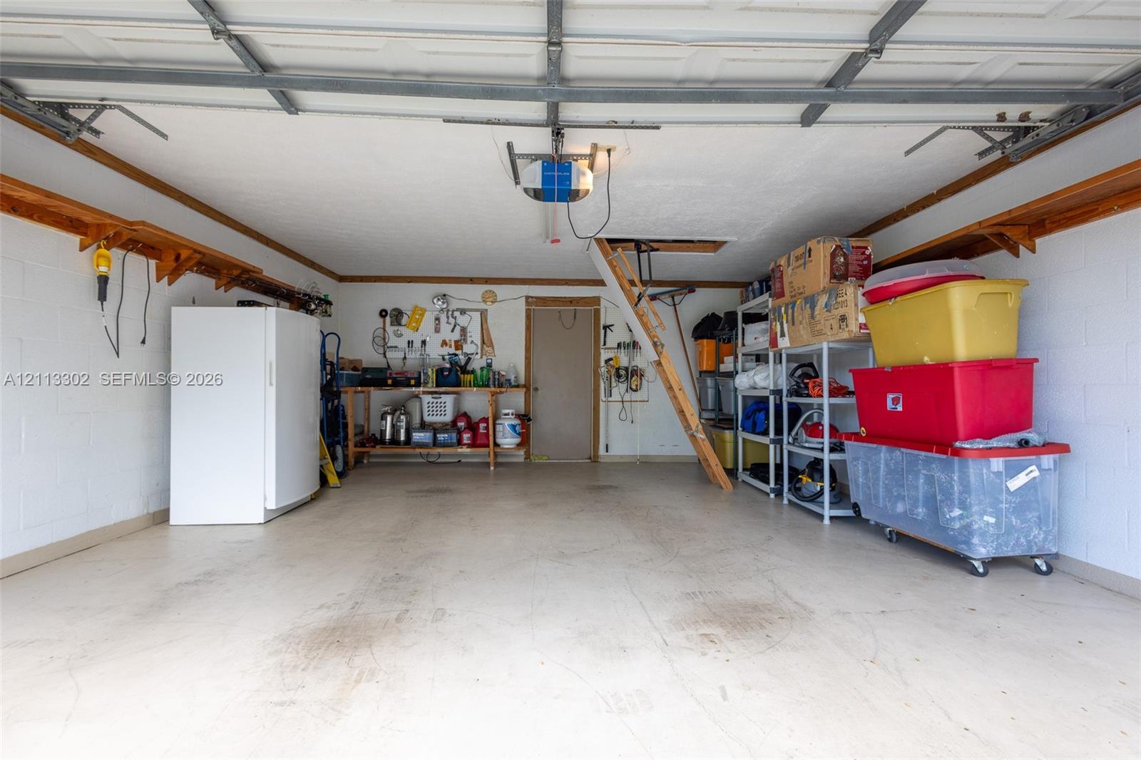Attached garage featuring concrete block walls, overhead door opener system, built-in wooden perimeter shelving, and pull-down attic access ladder
