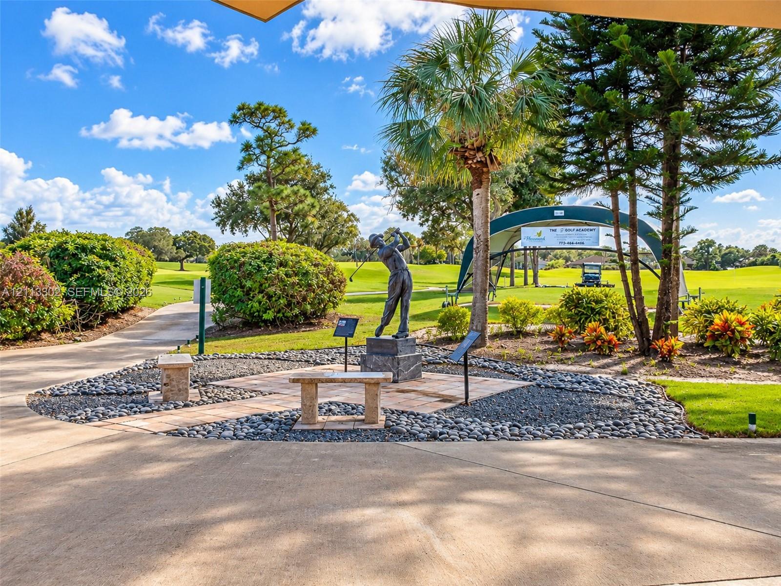 Landscaped grounds featuring stone benches, a central bronze statue, paved pathways, mature pine and palm trees, and golf course frontage