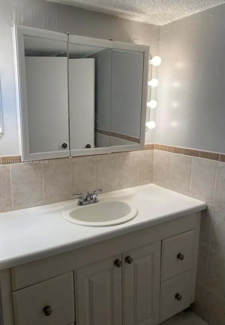 Vanity featuring built-in cabinetry with drawers, solid surface countertop, integrated basin, tri-fold mirrored medicine cabinet, and vertical lighting fixtures, accented by tile wainscoting