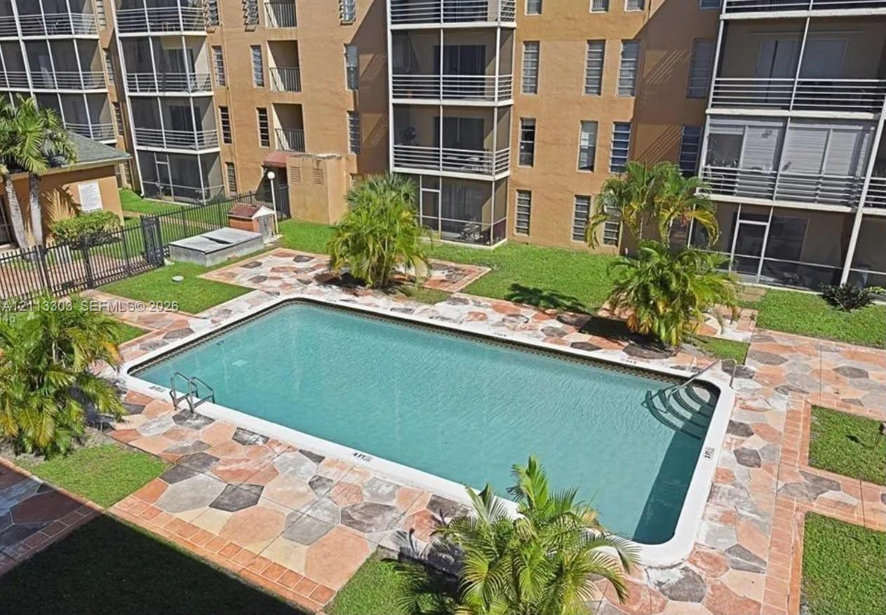 Rectangular swimming pool with metal ladder and steps, surrounded by multi-colored stone paver decking, manicured lawn, and mature palm trees
