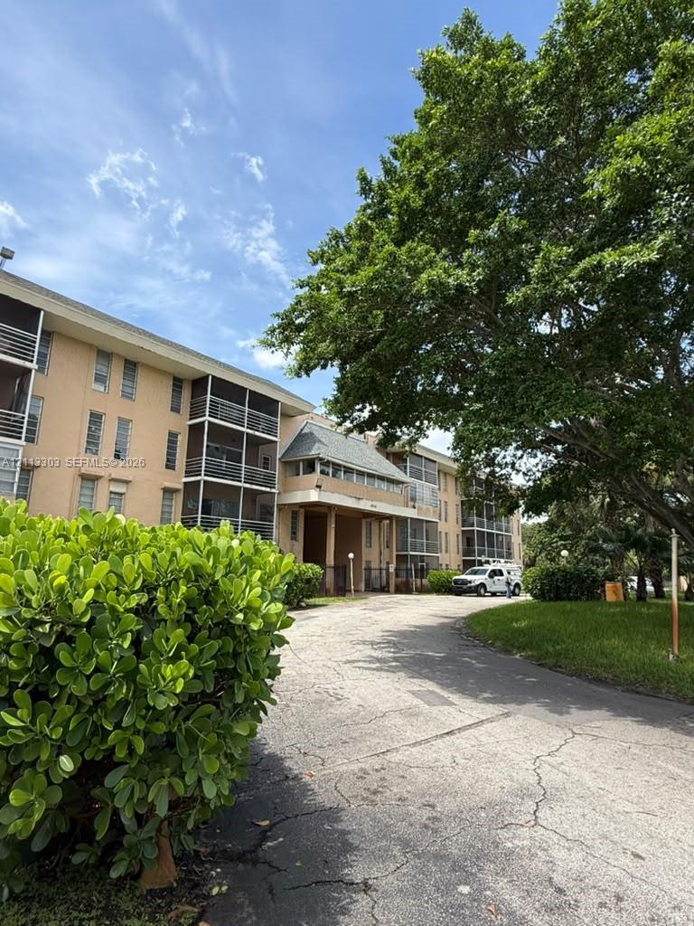 Multi-story building exterior featuring screened balconies, stucco facade, mature trees, and landscaped grounds