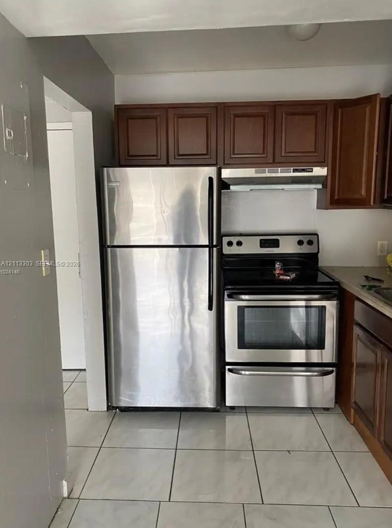 Kitchen featuring wood cabinetry, stainless steel refrigerator, stainless steel electric range, range hood, and tile flooring