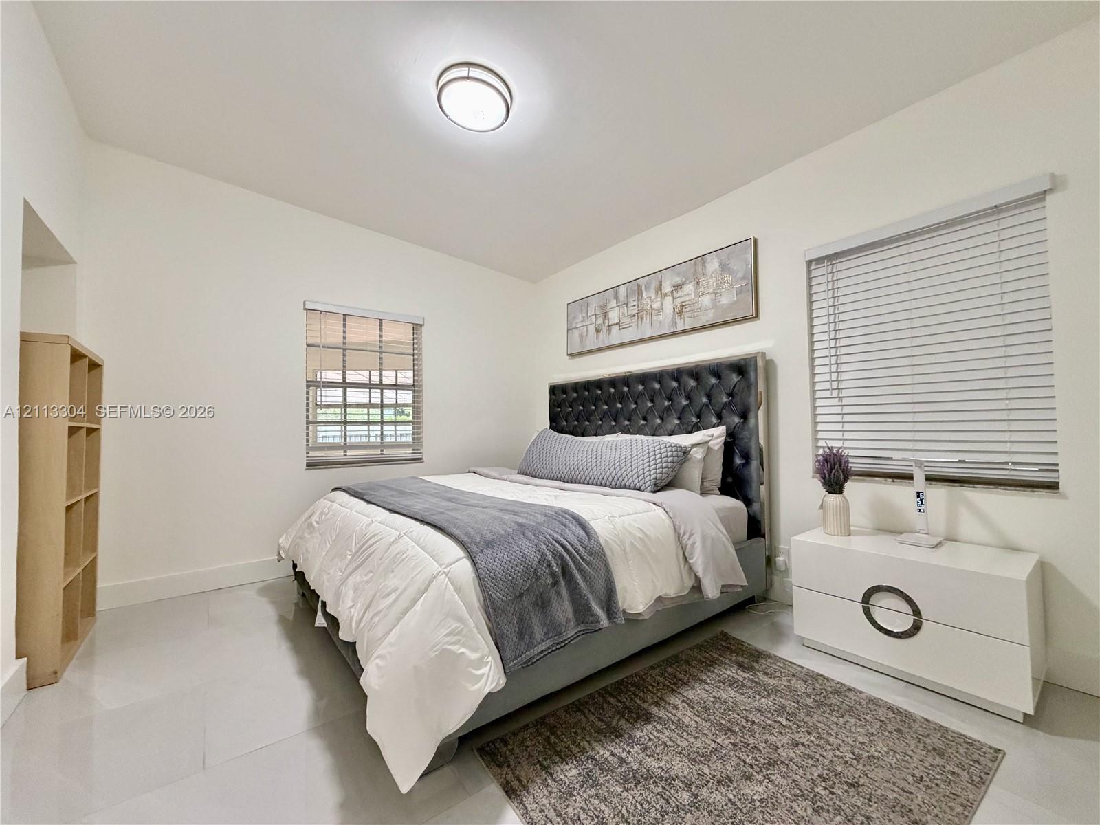 Neutral-toned interior featuring large-format tile flooring, recessed ceiling lighting, two windows with horizontal blinds, and a built-in open-shelving unit