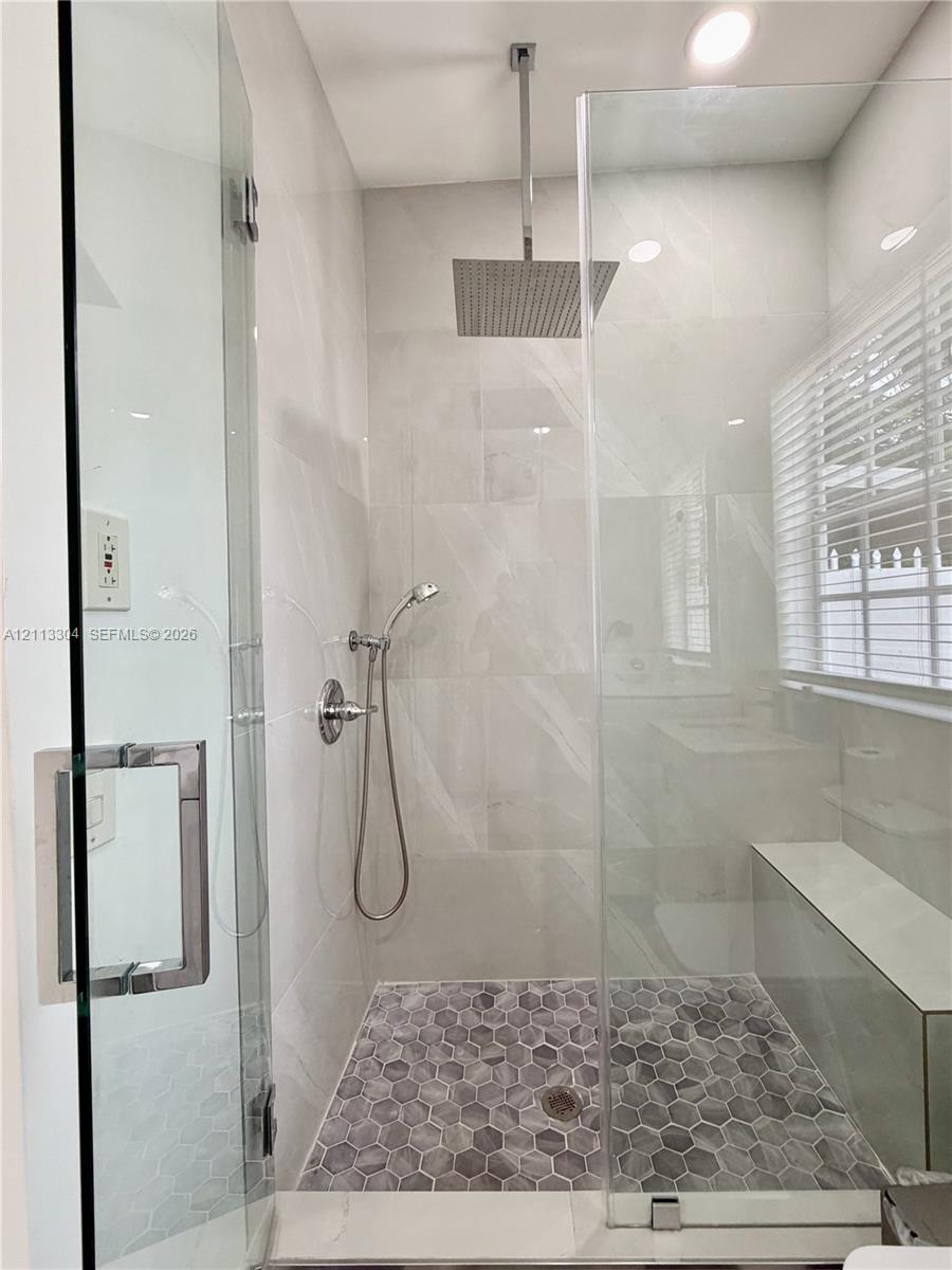 Walk-in shower featuring a frameless glass enclosure, ceiling-mounted rainfall showerhead, handheld sprayer, built-in bench, and grey hexagonal floor tile