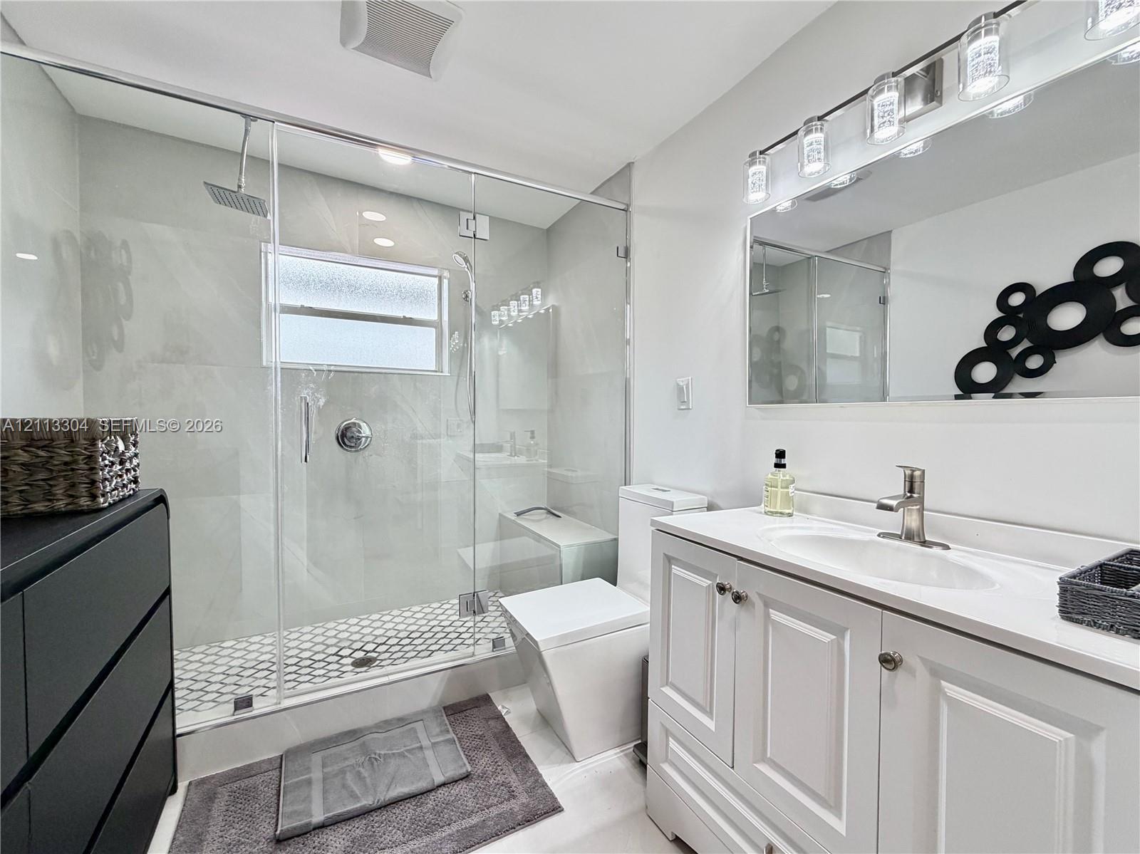Bathroom featuring a frameless glass-enclosed shower with large-format wall tile and hexagonal floor tile, a white vanity cabinet with an integrated sink, and a multi-bulb light fixture above a wide mirror