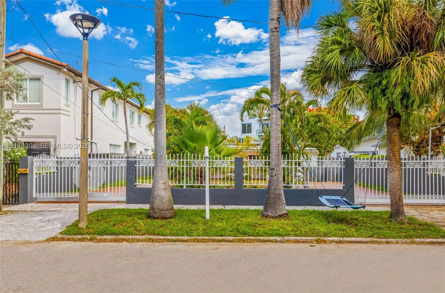 Exterior street-level view featuring a decorative metal perimeter fence with masonry pillars, manicured grass area, mature palm trees, and exterior post lighting