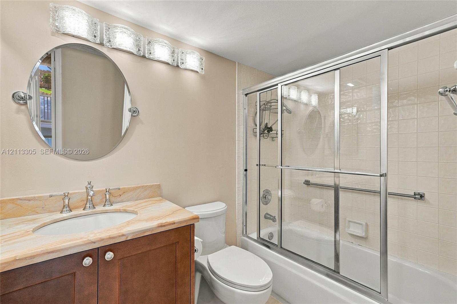 Bathroom featuring a wood-finish vanity with a stone-top integrated sink, circular mirror, multi-bulb vanity light, and a tub-shower combination with sliding glass doors