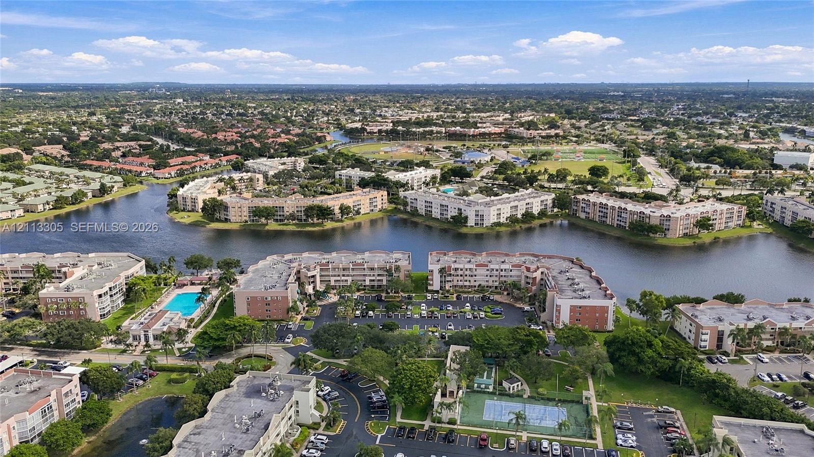 Aerial perspective of a multi-building residential complex featuring waterfront positioning, a swimming pool, a tennis court, and paved surface parking