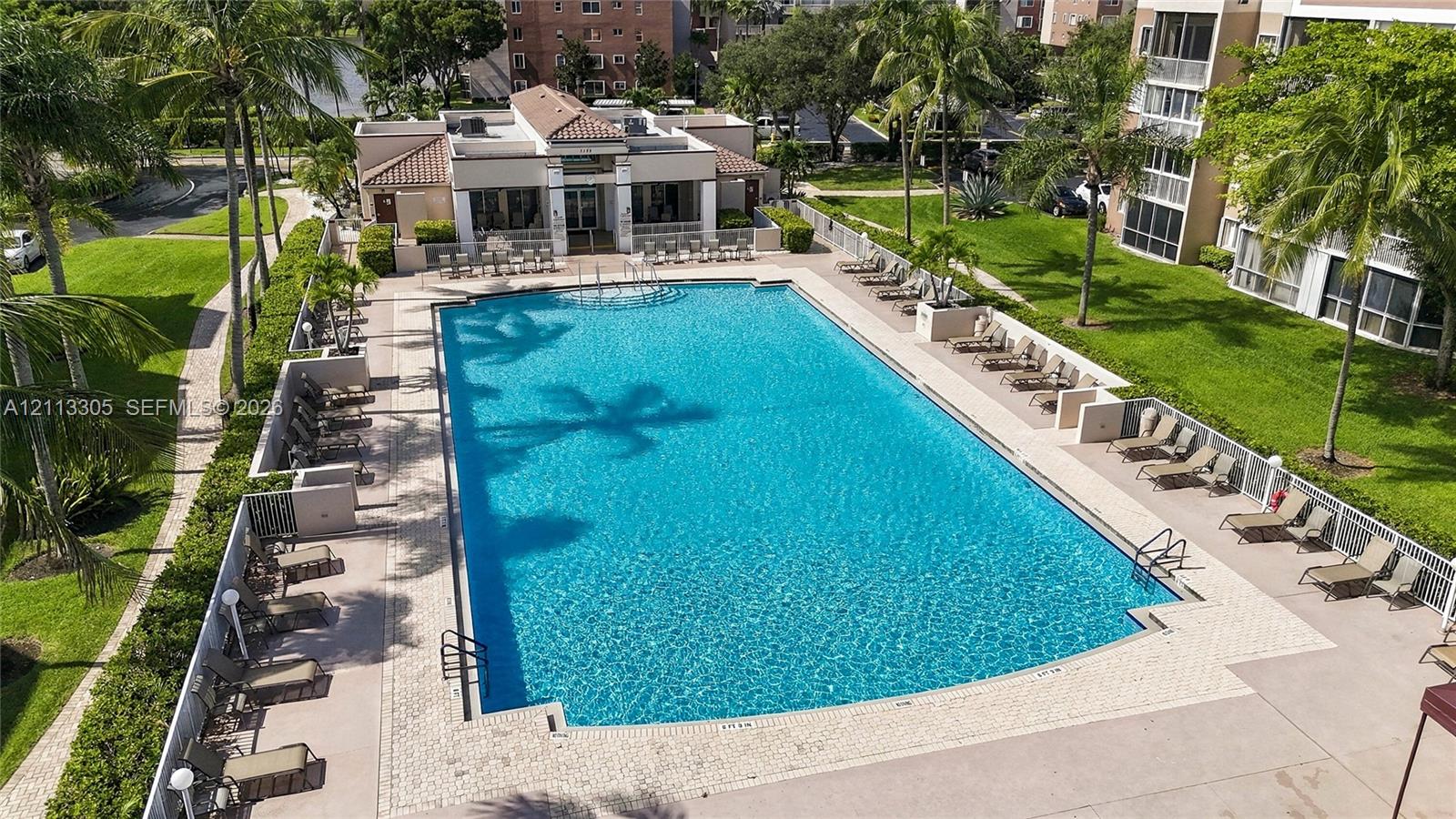 Community swimming pool featuring a concrete deck, paved border, and integrated pool entry steps, surrounded by manicured grounds and established palm trees