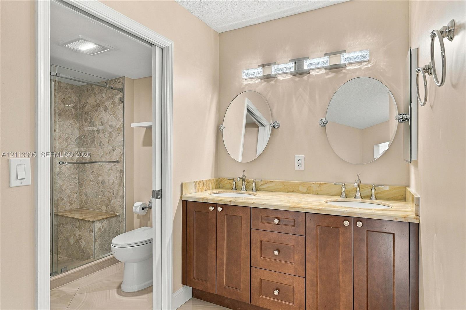 Double vanity featuring a stone countertop, under-mount sinks, wood cabinetry, dual oval wall-mounted mirrors, and an integrated glass-enclosed shower with tile surround