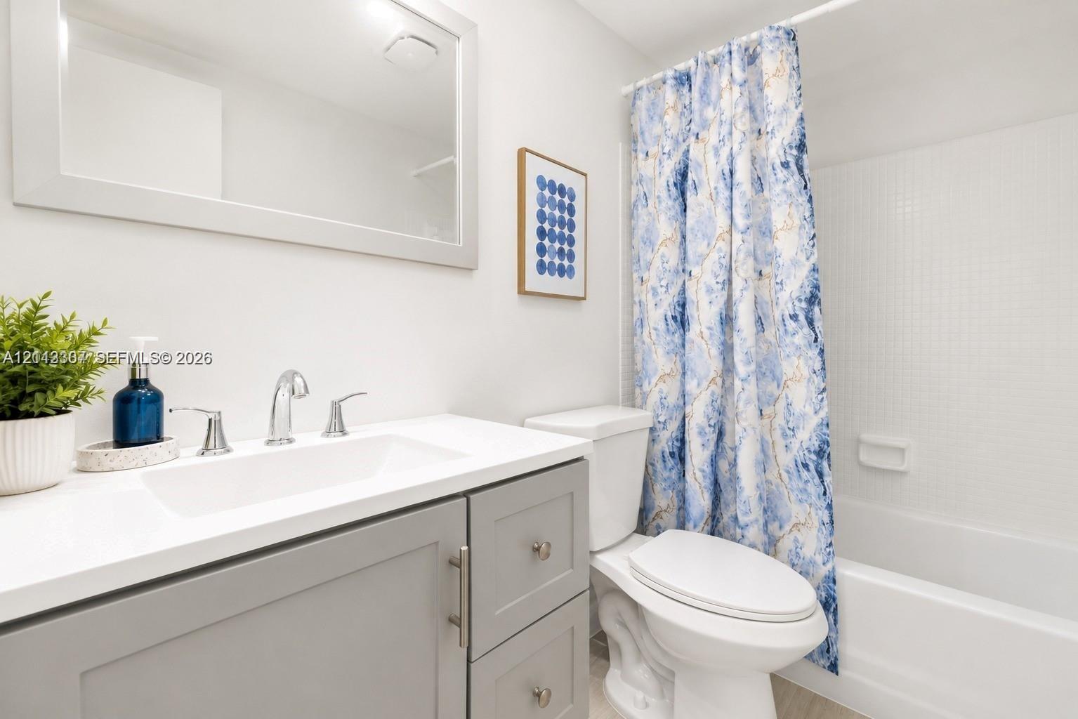 Bathroom featuring a grey shaker-style vanity, white countertop with an integrated sink, polished chrome faucet, framed wall mirror, and a tub-shower combination with white tiled surround Bathroom featuring a grey shaker-style vanity, white countertop with an integrated sink, polished chrome faucet, framed wall mirror, and a tub-shower combination with white tiled surround
