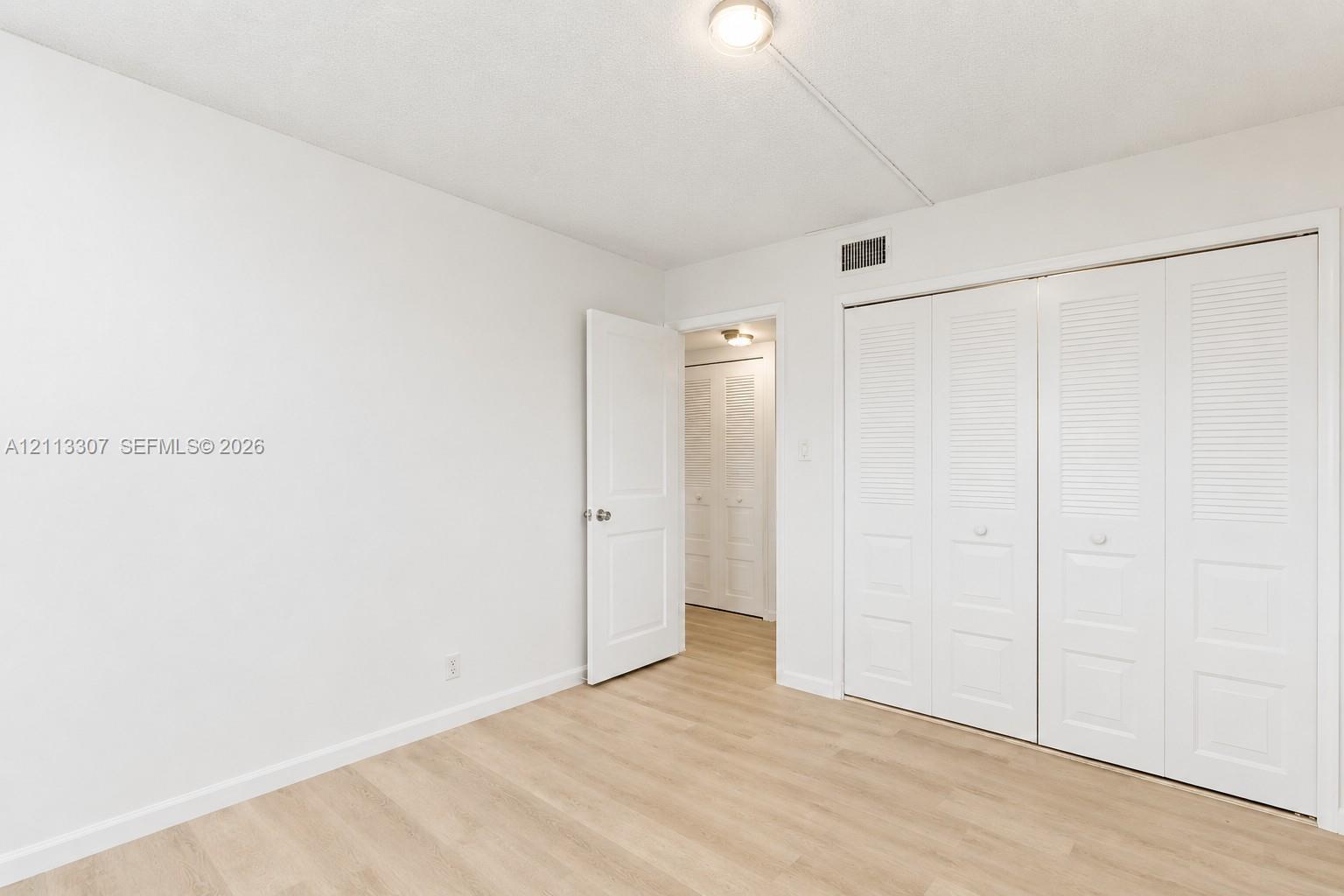 Neutral interior space featuring wood-finish flooring, white walls, flush-mount ceiling lighting, and built-in closet storage with louvered doors Neutral interior space featuring wood-finish flooring, white walls, flush-mount ceiling lighting, and built-in closet storage with louvered doors