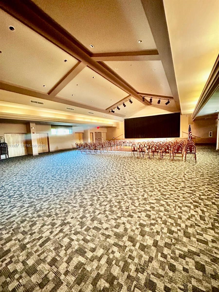 Expansive hall featuring high, multi-tiered tray ceilings with exposed beams, integrated recessed lighting, track lighting, and patterned wall-to-wall carpeting