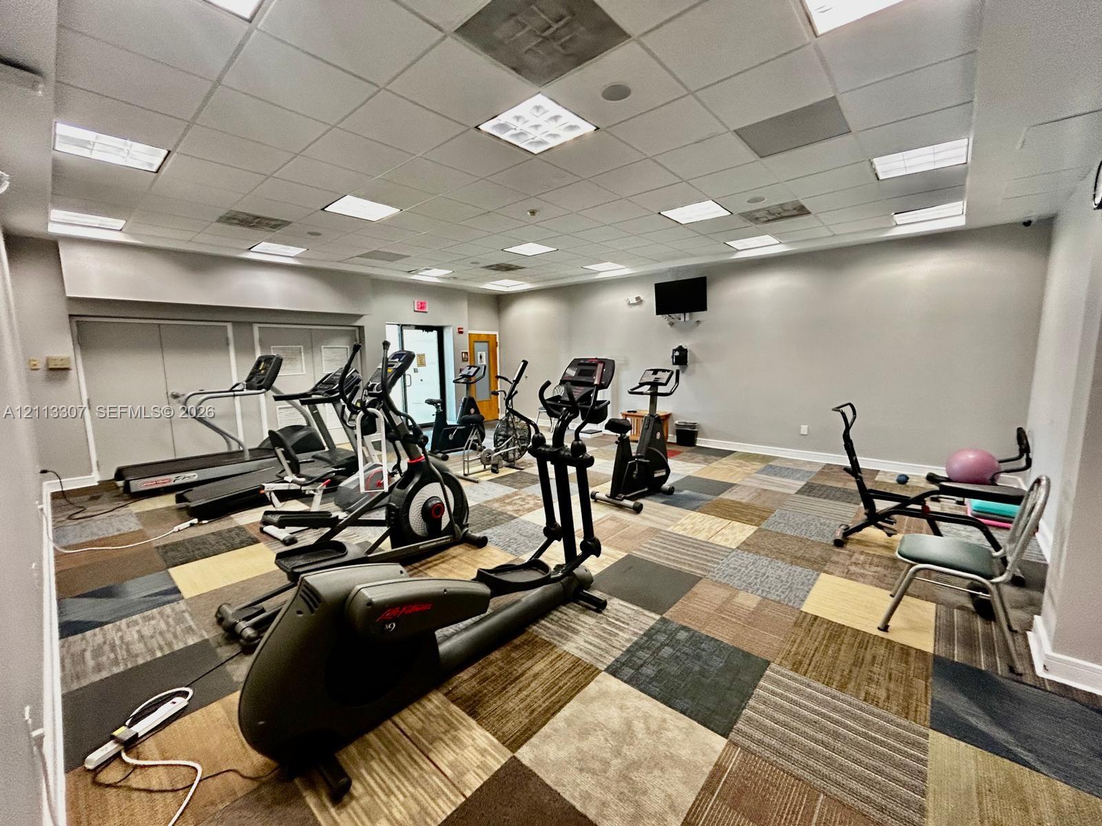 Fitness center featuring suspended grid ceiling, recessed panel lighting, and multi-toned modular carpet tiles