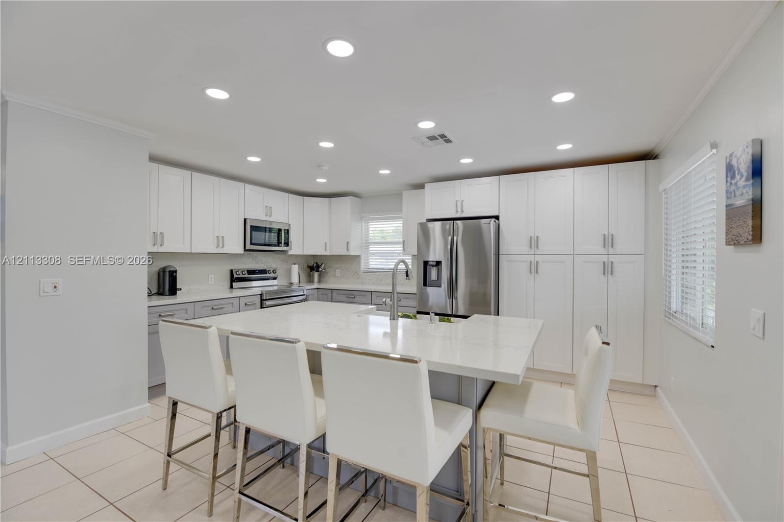 Kitchen featuring white cabinetry, an expansive central island with quartz countertops, stainless steel appliances, recessed ceiling lighting, and ceramic tile flooring Kitchen featuring white cabinetry, an expansive central island with quartz countertops, stainless steel appliances, recessed ceiling lighting, and ceramic tile flooring