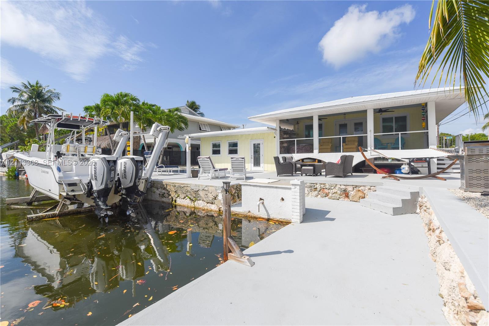 Waterfront property featuring a concrete seawall, boat lift, paved patio, and a covered porch with glass railing