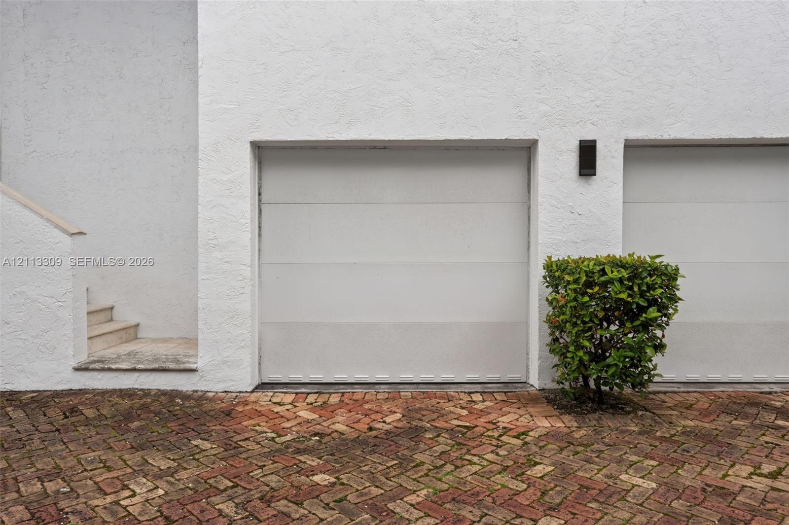 Multi-car garage featuring white paneled doors, textured stucco exterior, exterior wall-mounted lighting, and a herringbone-patterned brick paver driveway