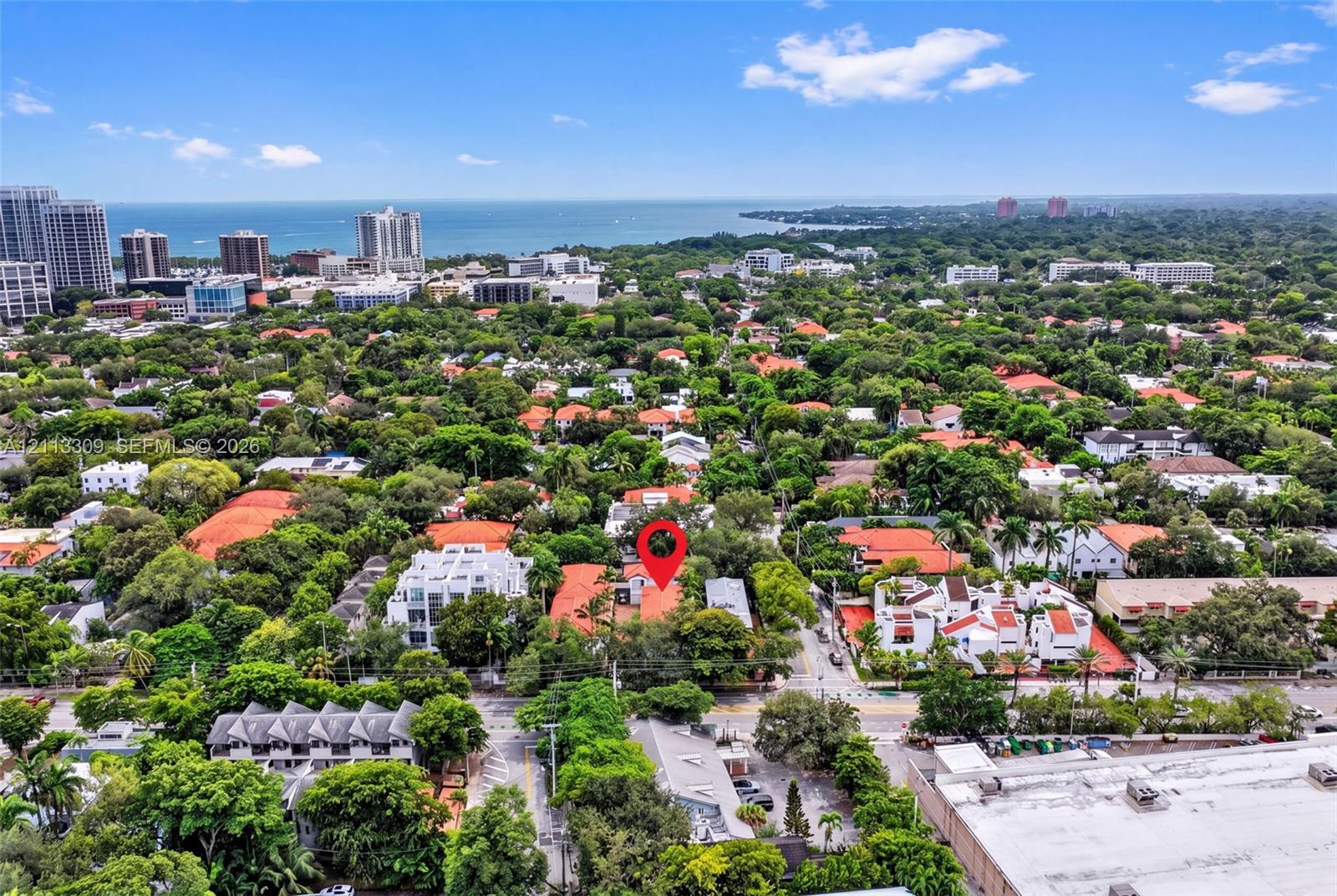 Aerial perspective featuring a red-tile roof structure situated within a densely wooded suburban setting, including prominent coastal and horizon scenery