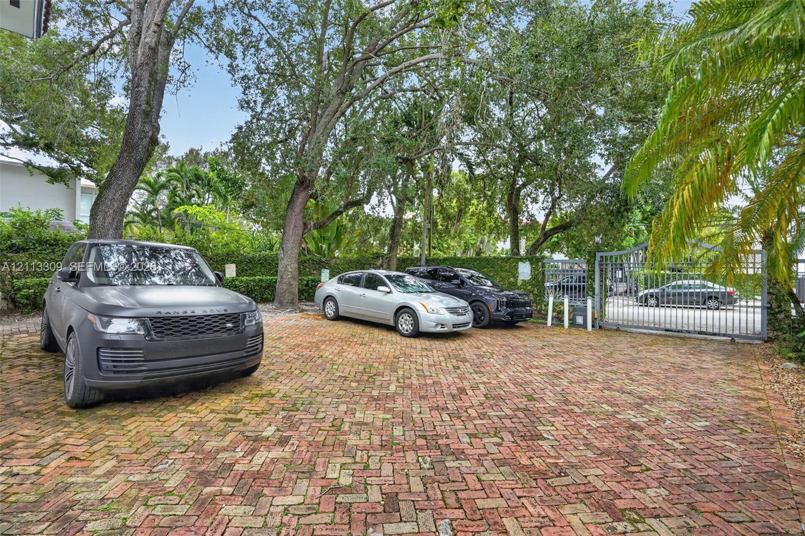 Brick-paved parking area featuring mature trees, perimeter hedging, and a metal security gate