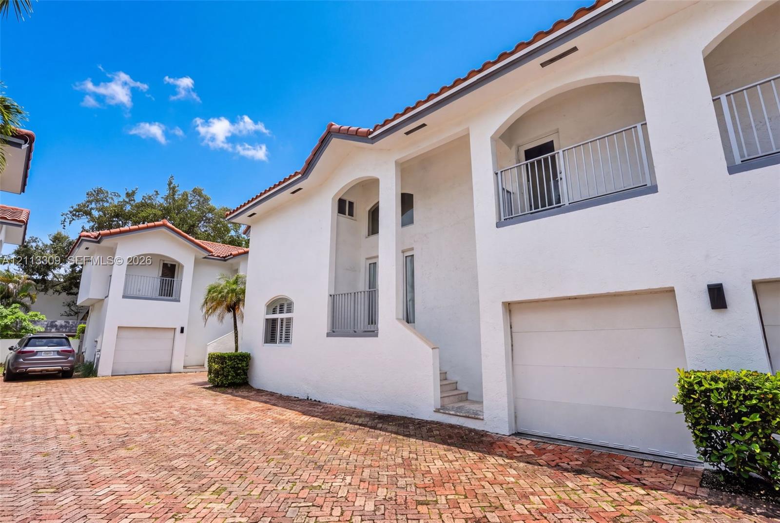 Stucco exterior featuring arched architectural detailing, multiple balconies with metal railings, a tiled roof, and a brick-paved driveway leading to enclosed garage spaces Stucco exterior featuring arched architectural detailing, multiple balconies with metal railings, a tiled roof, and a brick-paved driveway leading to enclosed garage spaces
