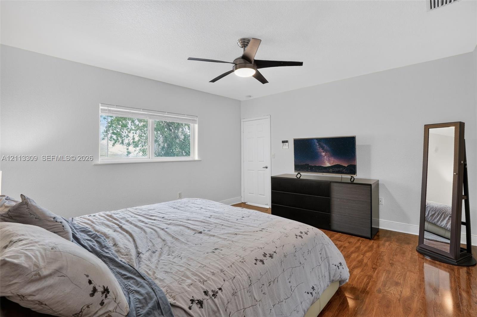 Wood-finish flooring, neutral wall tones, multi-blade ceiling fan with integrated lighting, and a window with horizontal blinds