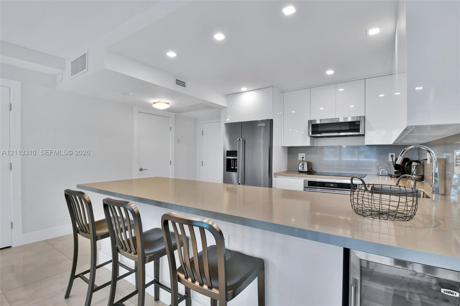 Kitchen featuring high-gloss white cabinetry, a neutral stone-surface peninsula, stainless steel appliances, recessed ceiling lighting, and tile flooring