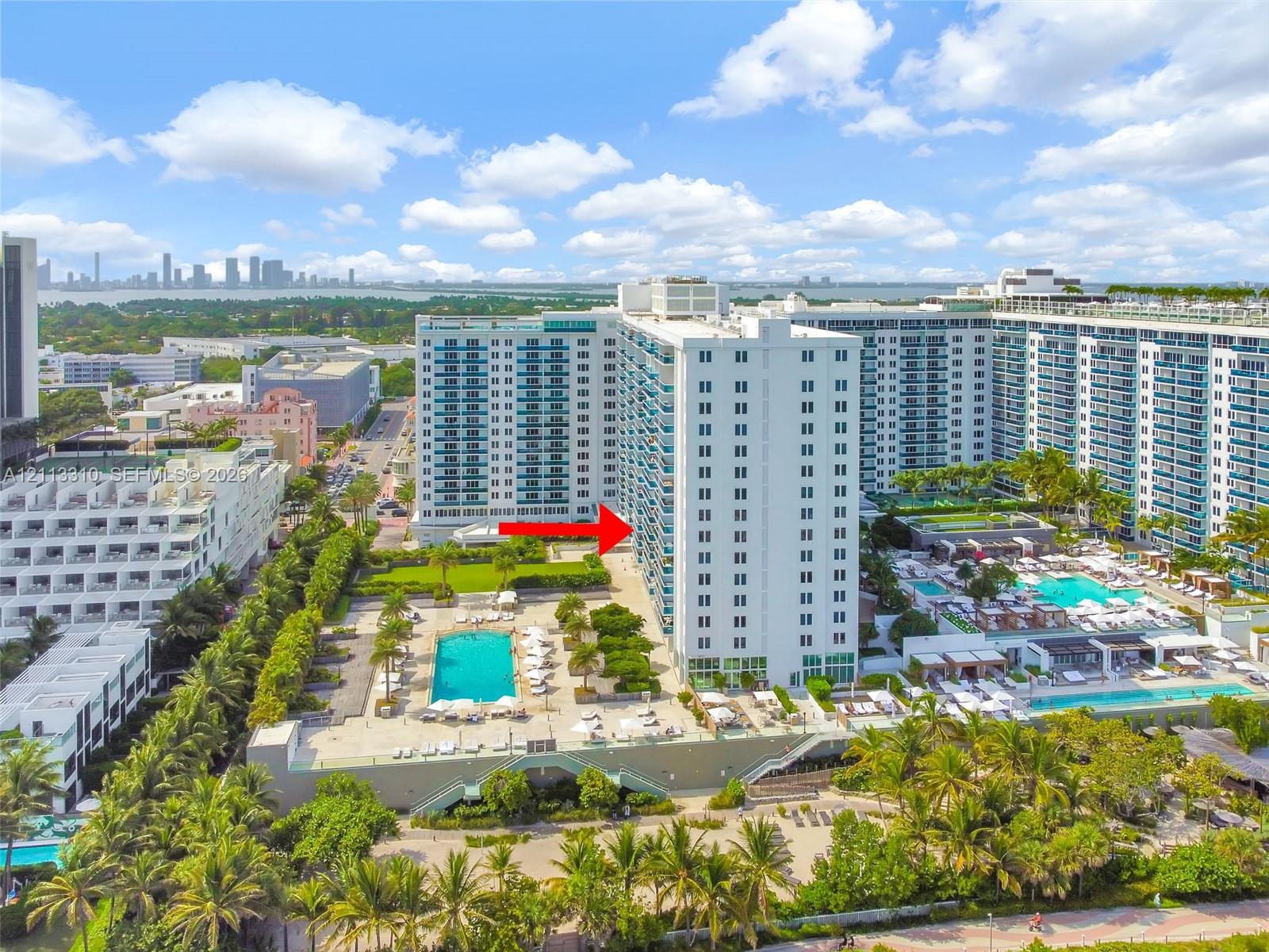 Aerial perspective of high-rise residential building featuring multiple swimming pools, expansive sun decks, manicured landscaping, and mature palm trees