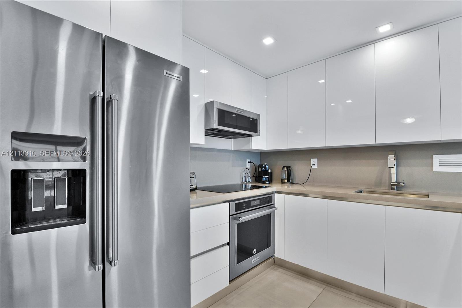 Kitchen featuring high-gloss white cabinetry, solid surface countertops, recessed lighting, and stainless steel appliances including a built-in oven, over-the-range microwave, and side-by-side refrigerator