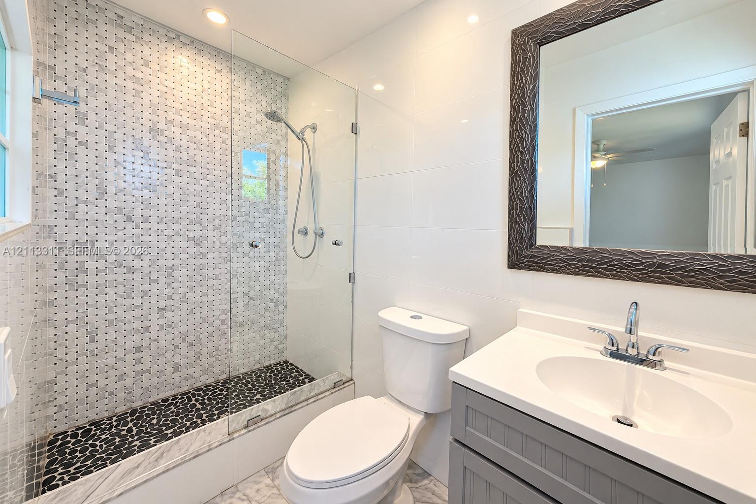 Walk-in shower featuring a frameless glass enclosure, decorative mosaic wall tile, and natural stone pebble flooring, complemented by a grey shaker-style vanity with a solid surface countertop