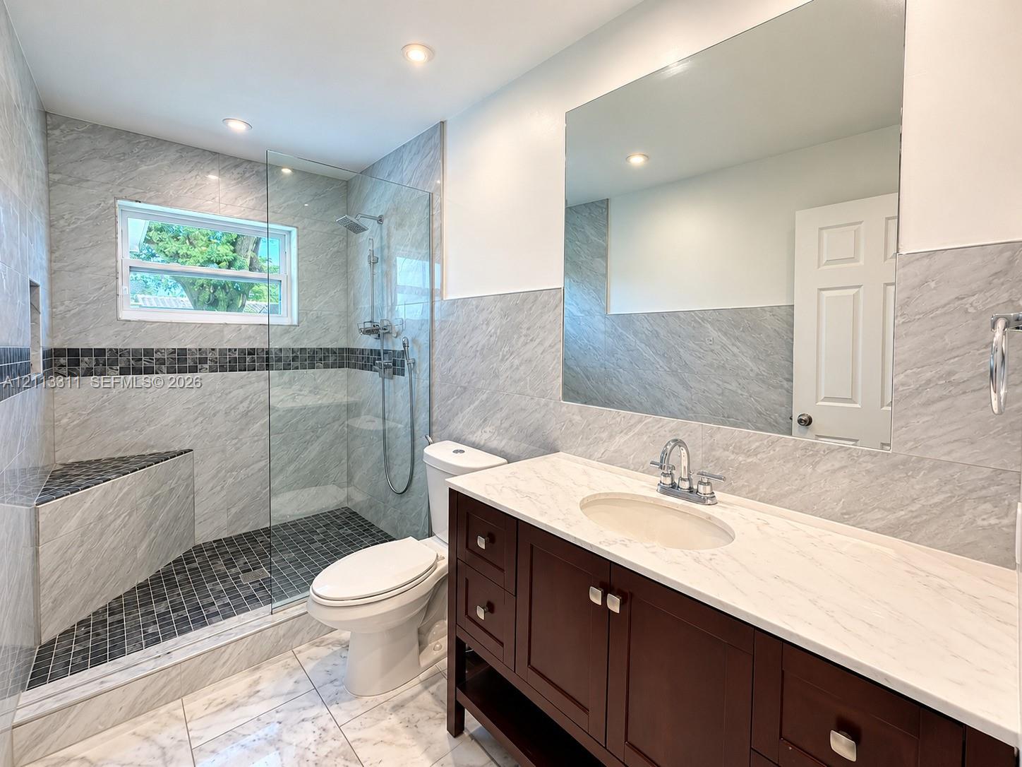Bathroom featuring a wood-finish vanity with a light-toned countertop, a walk-in shower with a glass panel and built-in bench, recessed lighting, and large-format wall tiling with a mosaic accent border